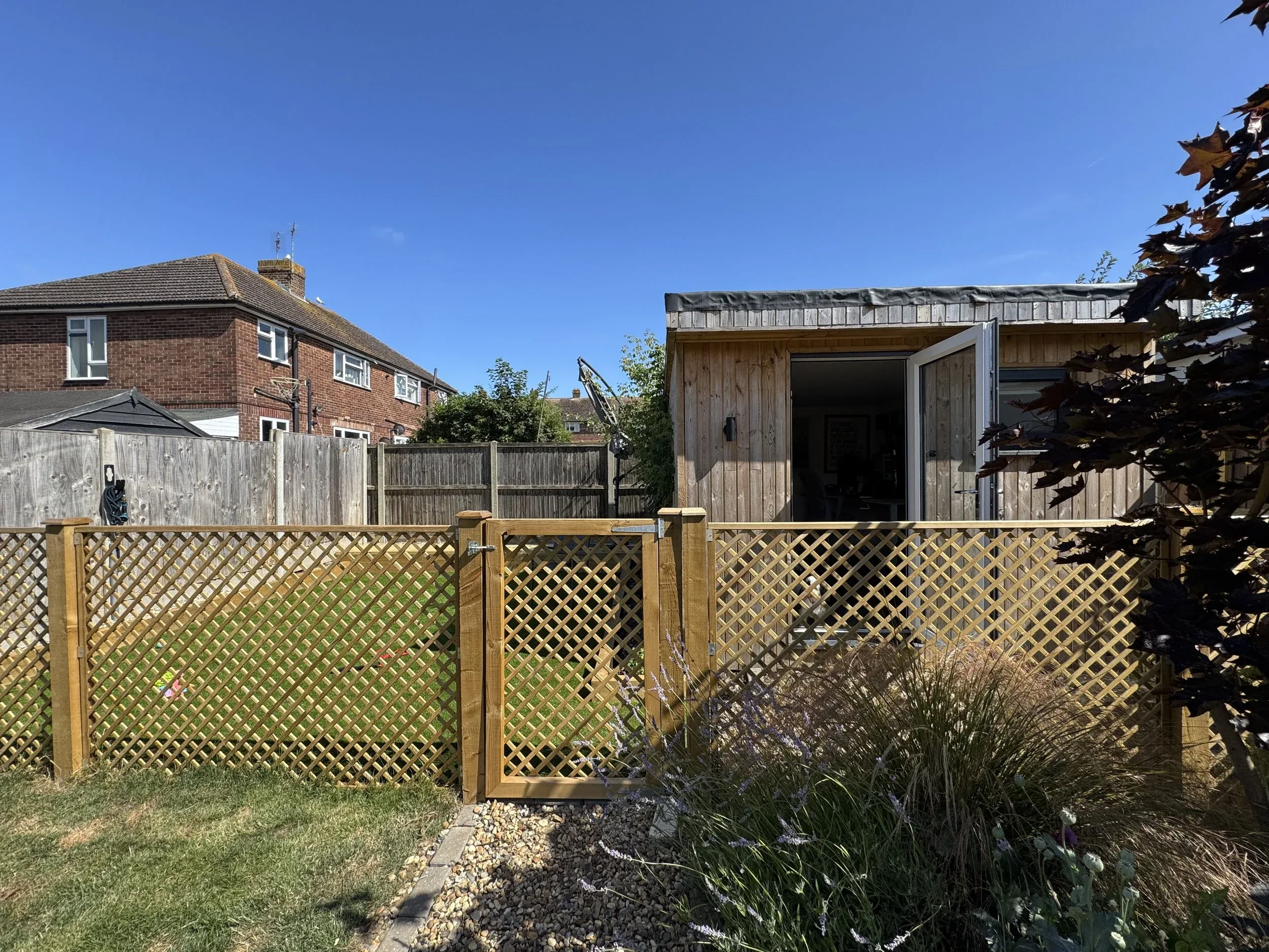 A small wooden garden shed with a glass door, situated behind a wooden lattice fence in a backyard.